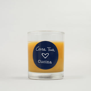 A clear glass candle with a yellow liquid inside, labeled 'CASA TUA CUCINA' with a heart symbol on the label.