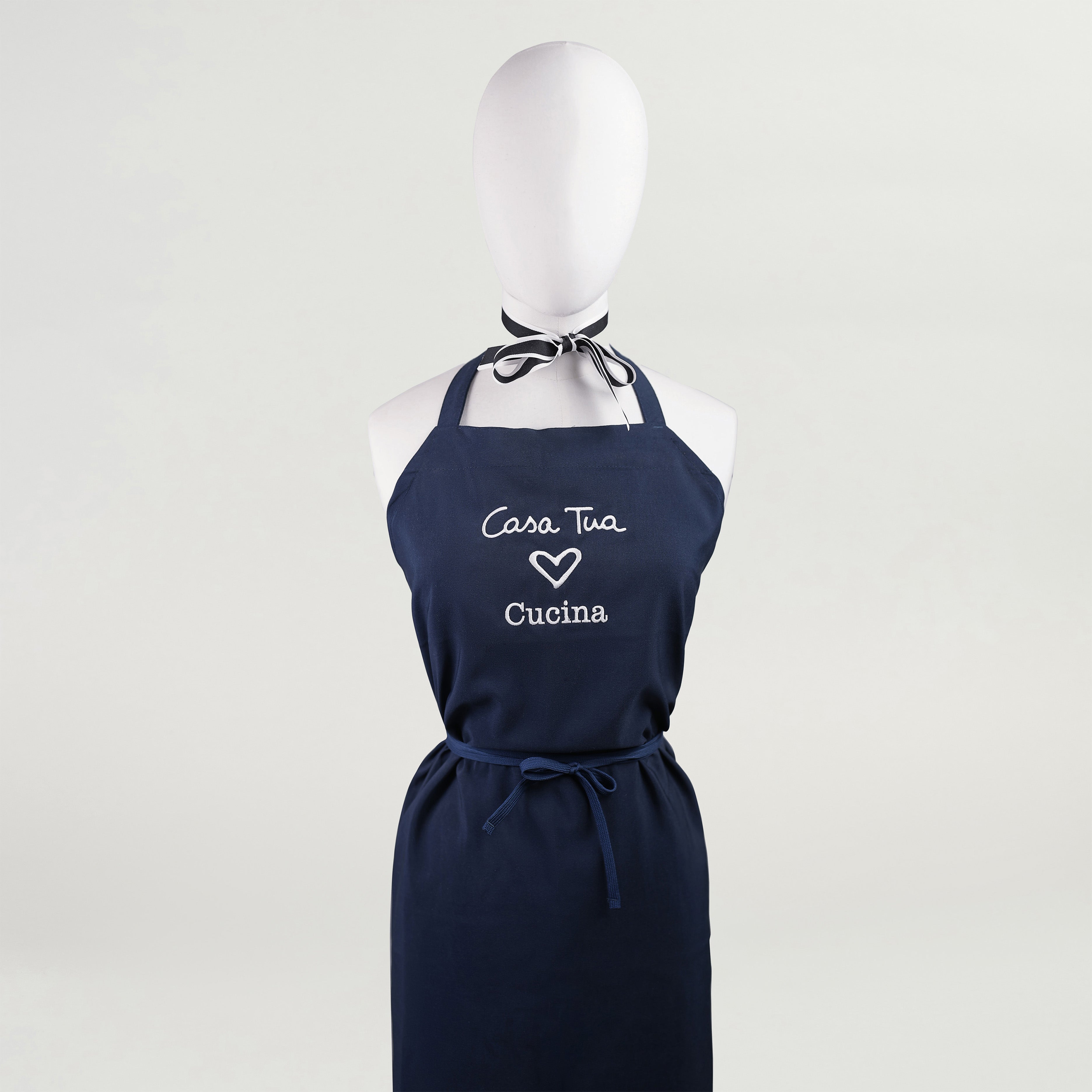 A navy blue kitchen apron with white text 'Casa Tua' and a heart symbol followed by 'Cucina' displayed on a mannequin.