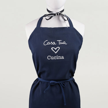 A navy blue kitchen apron with white text 'Casa Tua' and a heart symbol followed by 'Cucina' displayed on a mannequin.