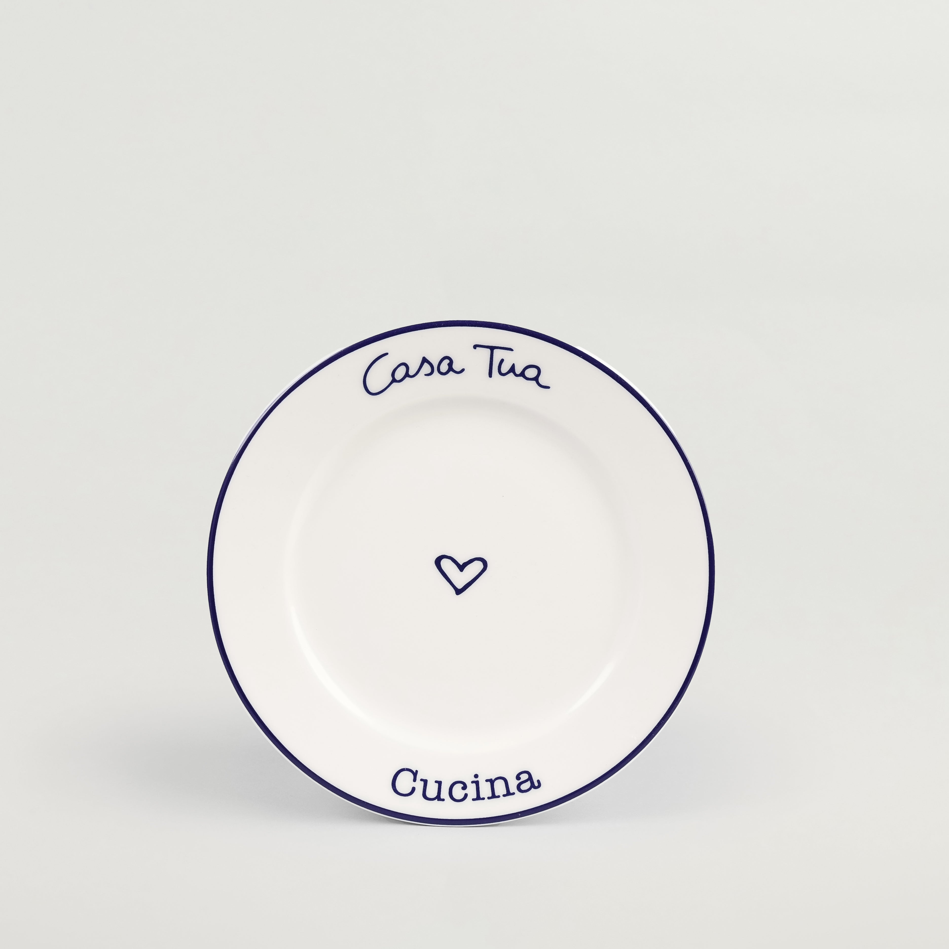 A white ceramic plate with blue rim and text reading 'Casa Tua Cucina' along with a heart symbol in the center.